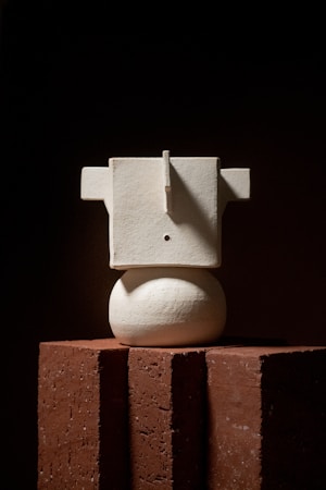 A minimalist sculpture composed of geometric shapes, including a rectangular form on top of an oval base. The sculpture is placed on reddish-brown bricks against a dark background. Strong lighting creates dramatic shadows and emphasizes the texture of the materials.