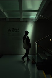 A person stands in a dimly lit space next to a wall with text indicating 'Museo de Sitio'. The environment appears modern and minimalist, with overhead beams and a reflective floor. The lighting casts a silhouette of the person, creating a shadowy effect.