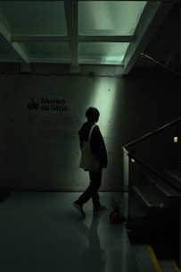A person stands in a dimly lit space next to a wall with text indicating 'Museo de Sitio'. The environment appears modern and minimalist, with overhead beams and a reflective floor. The lighting casts a silhouette of the person, creating a shadowy effect.