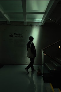 A person stands in a dimly lit space next to a wall with text indicating 'Museo de Sitio'. The environment appears modern and minimalist, with overhead beams and a reflective floor. The lighting casts a silhouette of the person, creating a shadowy effect.