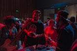 A group of people gathered in a dimly lit room, possibly participating in a music-related event. One individual is holding a guitar, another appears to be interacting with an electronic device on a table, while others are engaged in conversation. The lighting casts a reddish hue over the scene, emphasizing the informal and engaging atmosphere.