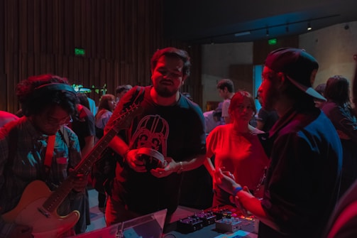 A group of people gathered in a dimly lit room, possibly participating in a music-related event. One individual is holding a guitar, another appears to be interacting with an electronic device on a table, while others are engaged in conversation. The lighting casts a reddish hue over the scene, emphasizing the informal and engaging atmosphere.