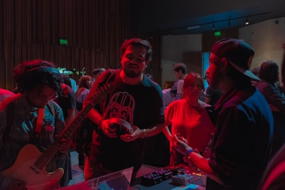 A group of people gathered in a dimly lit room, possibly participating in a music-related event. One individual is holding a guitar, another appears to be interacting with an electronic device on a table, while others are engaged in conversation. The lighting casts a reddish hue over the scene, emphasizing the informal and engaging atmosphere.