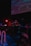 A DJ is performing at a dimly lit venue with a large screen displaying visuals in the background. The setting includes technical equipment and a small 'ON AIR' sign illuminated in red.