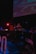 A DJ is performing at a dimly lit venue with a large screen displaying visuals in the background. The setting includes technical equipment and a small 'ON AIR' sign illuminated in red.