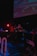 A DJ is performing at a dimly lit venue with a large screen displaying visuals in the background. The setting includes technical equipment and a small 'ON AIR' sign illuminated in red.