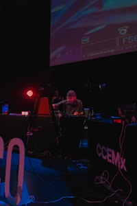 A DJ is performing at a dimly lit venue with a large screen displaying visuals in the background. The setting includes technical equipment and a small 'ON AIR' sign illuminated in red.