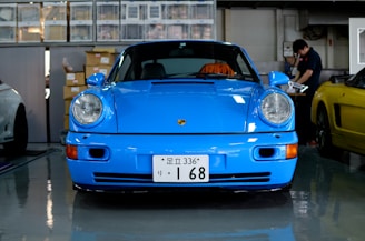 A bright blue sports car is parked indoors in a garage or workshop setting. A person is seen working in the background near a yellow car. Various boxes and automotive parts are stacked behind the blue car.