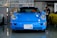 A bright blue sports car is parked indoors in a garage or workshop setting. A person is seen working in the background near a yellow car. Various boxes and automotive parts are stacked behind the blue car.