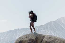 A person stands on a large rock, looking out through binoculars. They are wearing athletic clothing and carrying a large backpack, suggesting they are hiking or exploring. In the background, there are mountains and a clear sky.