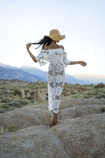 a woman in a white dress and hat walking on a rock