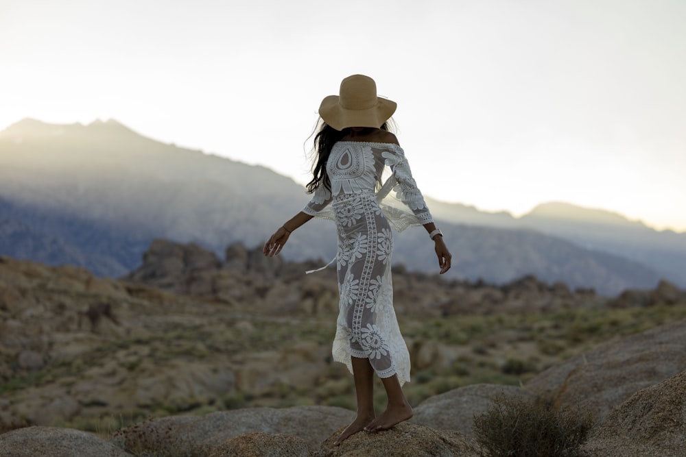 a woman in a white dress and hat standing on a rock