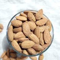 A glass bowl filled with whole almonds is placed on a light-colored surface. The almonds have a natural brown, slightly textured shell.
