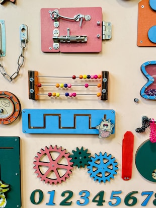 A sensory board featuring various tactile objects and elements such as a latch, an abacus with colorful beads, gears, and numbers. The board is designed with interactive components in different bright colors like red, blue, and orange.