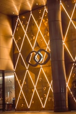An elegant, geometric wall display with golden lighting and triangular patterns features prominently. Intricate, illuminated lines criss-cross the surface, creating a diamond-like pattern. A central abstract logo or emblem stands out amidst the design. A large column is visible on the right, and glass doors on the left reveal a glimpse of a decorated interior.