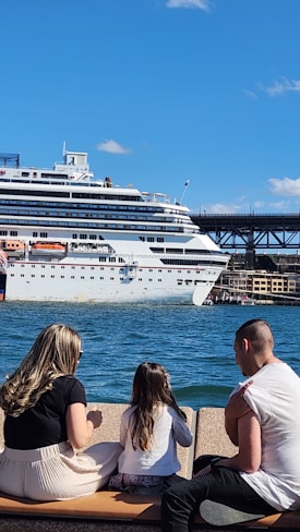 A family of three, consisting of two adults and a child, is seated on a bench overlooking a large cruise ship docked in a harbor. The scene is set under a clear blue sky. The woman has long hair and is wearing a light skirt, while the man has short hair, and the child has long hair as well.