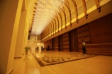 An opulent interior features a long hallway with a high ceiling adorned with intricate, glowing light fixtures. The floor is polished and reflective, displaying ornate patterns. Wooden paneling lines one side, complemented by large, intricately designed doors.