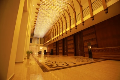 An opulent interior features a long hallway with a high ceiling adorned with intricate, glowing light fixtures. The floor is polished and reflective, displaying ornate patterns. Wooden paneling lines one side, complemented by large, intricately designed doors.