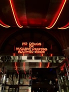 A neon sign with red light stating 'No drugs or nuclear weapons allowed inside' is mounted above a metallic entrance. The environment suggests an urban setting with reflective surfaces and a glimpse into an interior space that contains various decorations.