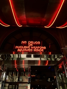 A neon sign with red light stating 'No drugs or nuclear weapons allowed inside' is mounted above a metallic entrance. The environment suggests an urban setting with reflective surfaces and a glimpse into an interior space that contains various decorations.