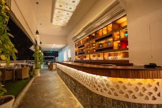 A stylish bar area features a long wooden counter with intricate latticework and glowing warm lighting underneath. Behind the counter, shelves are illuminated, showcasing various colored glasses. The ceiling is adorned with decorative light panels, and several pendant lights hang over the space. To the left, potted plants and dining tables with chairs are arranged, with an open view to a nighttime setting.
