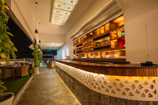 A stylish bar area features a long wooden counter with intricate latticework and glowing warm lighting underneath. Behind the counter, shelves are illuminated, showcasing various colored glasses. The ceiling is adorned with decorative light panels, and several pendant lights hang over the space. To the left, potted plants and dining tables with chairs are arranged, with an open view to a nighttime setting.