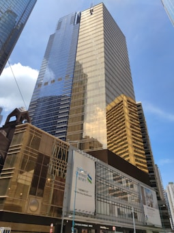 A towering modern skyscraper with reflective glass surfaces stands prominently under a clear blue sky. The building features a combination of blue and gold hues, with intricate architectural designs. In the foreground, a large building with a Standard Chartered Bank sign is visible.