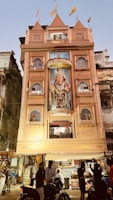 A multi-story building with an ornate facade featuring a large statue of Ganesha, adorned with flags on the roof. The building is lit up and has people gathered in front, some on motorcycles. The surroundings include other older structures and signs of a bustling street atmosphere.