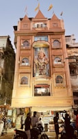 A multi-story building with an ornate facade featuring a large statue of Ganesha, adorned with flags on the roof. The building is lit up and has people gathered in front, some on motorcycles. The surroundings include other older structures and signs of a bustling street atmosphere.