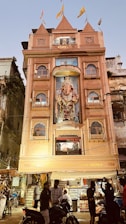 A multi-story building with an ornate facade featuring a large statue of Ganesha, adorned with flags on the roof. The building is lit up and has people gathered in front, some on motorcycles. The surroundings include other older structures and signs of a bustling street atmosphere.