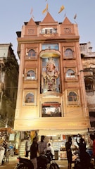 A multi-story building with an ornate facade featuring a large statue of Ganesha, adorned with flags on the roof. The building is lit up and has people gathered in front, some on motorcycles. The surroundings include other older structures and signs of a bustling street atmosphere.