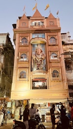A multi-story building with an ornate facade featuring a large statue of Ganesha, adorned with flags on the roof. The building is lit up and has people gathered in front, some on motorcycles. The surroundings include other older structures and signs of a bustling street atmosphere.