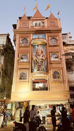 A multi-story building with an ornate facade featuring a large statue of Ganesha, adorned with flags on the roof. The building is lit up and has people gathered in front, some on motorcycles. The surroundings include other older structures and signs of a bustling street atmosphere.