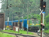 A green and yellow electric locomotive is hauling a blue freight train through lush greenery. Overhead electric lines and multiple utility poles run along the railway track. A red traffic signal indicating stop is visible to the right, along with a sign displaying directions.