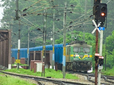 A green and yellow electric locomotive is hauling a blue freight train through lush greenery. Overhead electric lines and multiple utility poles run along the railway track. A red traffic signal indicating stop is visible to the right, along with a sign displaying directions.