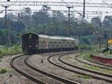 A long passenger train is moving along curved railway tracks surrounded by lush green vegetation. Overhead, multiple cables and electric lines stretch across the scene, indicating electrified railway lines. The environment is open, with trees in the distance and a slightly cloudy sky. A train signal light can be seen, and a station sign with some text is partially visible on the right.
