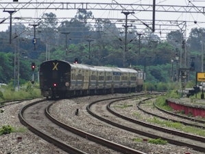 A long passenger train is moving along curved railway tracks surrounded by lush green vegetation. Overhead, multiple cables and electric lines stretch across the scene, indicating electrified railway lines. The environment is open, with trees in the distance and a slightly cloudy sky. A train signal light can be seen, and a station sign with some text is partially visible on the right.