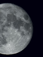 A close-up view of the moon, showing detailed textures, craters, and dark patches on its surface. The lighting highlights these features, creating a stark contrast against the blackness of space.