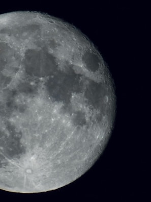A close-up view of the moon, showing detailed textures, craters, and dark patches on its surface. The lighting highlights these features, creating a stark contrast against the blackness of space.
