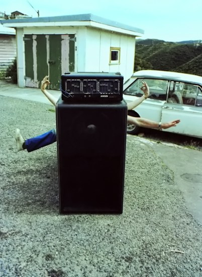 A large amplifier or speaker is positioned in the center of a gravel area. It appears as if disembodied arms and legs are playfully sticking out from behind the speaker. Behind this setup, there is an old white car and a small building with a green and white door. The image appears to have been taken outdoors in a hilly or mountainous area.