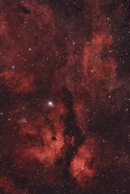 A deep space scene with red nebulae and clusters of stars scattered throughout. Dark patches and bright spots create a vivid contrast against the glowing gas clouds.