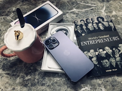 A sleek smartphone rests on its box, with charging cables neatly coiled nearby. Adjacent to the phone is a book titled 'World's Greatest Entrepreneurs,' featuring black and white portraits on the cover. A pink mug with a unique lid adorned with a gold horse head is placed nearby, all set on a marble surface.