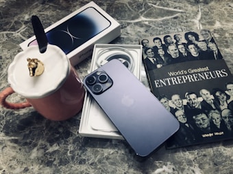 A sleek smartphone rests on its box, with charging cables neatly coiled nearby. Adjacent to the phone is a book titled 'World's Greatest Entrepreneurs,' featuring black and white portraits on the cover. A pink mug with a unique lid adorned with a gold horse head is placed nearby, all set on a marble surface.