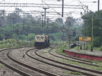 A train is approaching along a set of railway tracks surrounded by greenery. Overhead power lines and signals are visible, and a platform with a station sign labeled 'DHANDERI' can be seen on the right.