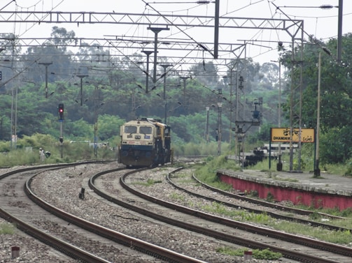 A train is approaching along a set of railway tracks surrounded by greenery. Overhead power lines and signals are visible, and a platform with a station sign labeled 'DHANDERI' can be seen on the right.