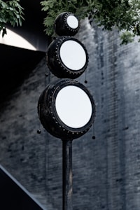 A vertical structure featuring three circular objects stacked in a totem-like formation. Each circular object is framed with dark material and features a white, reflective surface. The backdrop consists of a blurred wall with a textured, gray appearance, and there are green leaves hanging from the top left corner.