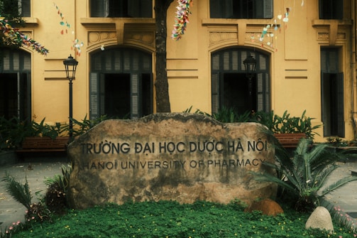 A large stone sign engraved with the name 'Hanoi University of Pharmacy' is set in front of a yellow building with arched windows. The area is surrounded by greenery, including ferns and other plants. Festive strings with colorful decorations hang overhead, and the atmosphere is complemented by two traditional-style street lamps.