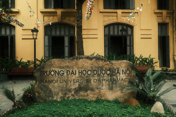 A large stone sign engraved with the name 'Hanoi University of Pharmacy' is set in front of a yellow building with arched windows. The area is surrounded by greenery, including ferns and other plants. Festive strings with colorful decorations hang overhead, and the atmosphere is complemented by two traditional-style street lamps.