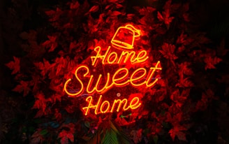 A neon sign in bright red-orange displays the words 'Home Sweet Home' against a backdrop of dark red leaves. The sign is the focal point amidst the textured and layered foliage.