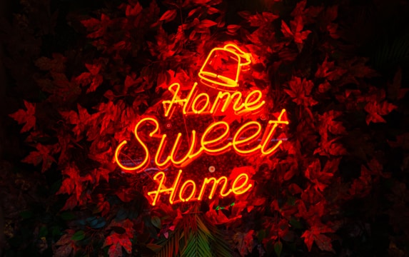 A neon sign in bright red-orange displays the words 'Home Sweet Home' against a backdrop of dark red leaves. The sign is the focal point amidst the textured and layered foliage.